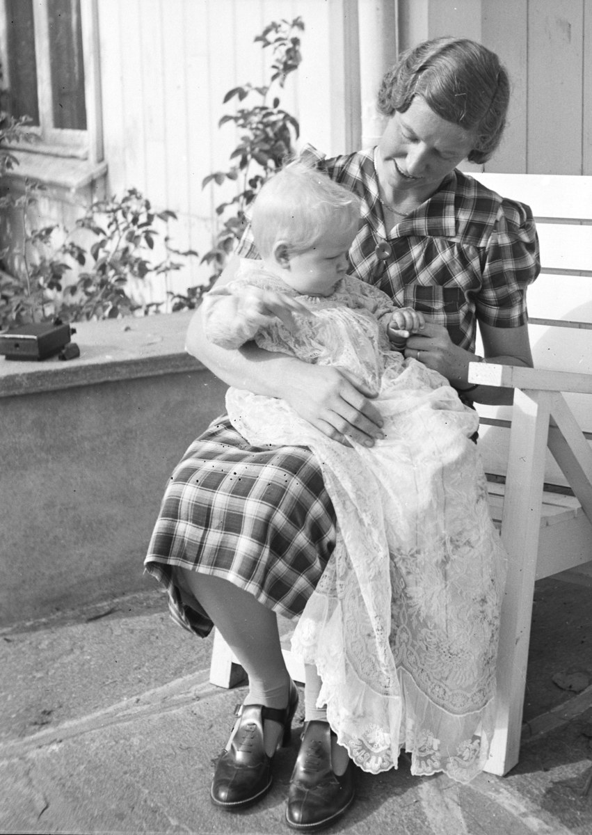 Eine Frau sitzt auf einer Bank und hält ein Baby, beide teilen einen warmen, liebevollen Moment in einem Raum mit üppigen Pflanzen und einem Fenster im Hintergrund.