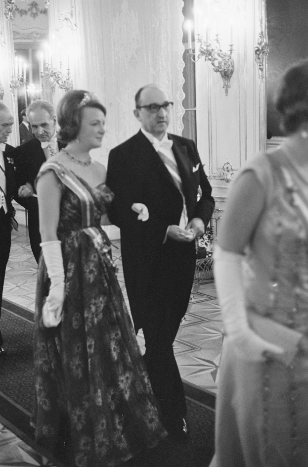 Ein Mann in einem schwarzen Blazer, weißem Hemd und schwarzen Hosen sowie eine Frau in einem eleganten Kleid gehen einen Flur entlang, umgeben von einer Gruppe von Menschen, die auf dem Boden stehen, mit ein paar an der Wand montierten Lampen, die eine gemütliche Atmosphäre schaffen.