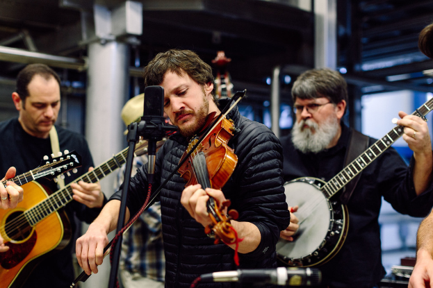 Eine Gruppe von Musikern in schwarzer Kleidung, die Instrumente spielen, mit einem Geigenvirtuosen vor einem Mikrofon und einem Gebäude im Hintergrund.