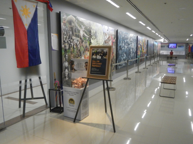 Langer Flur mit einer philippinischen Flagge an der Wand, Bildtafeln mit Text und Bildern, Ribbon-Ständer, Bänke, Deckenleuchten und ein Bildschirm mit Museums-Informationen im Hintergrund.