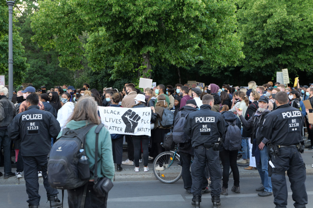 Eine große Gruppe von Menschen nimmt an einer Black Lives Matter Demonstration in Berlin teil, einige halten Schilder und andere tragen Kappen und Taschen, vorne steht ein Fahrrad und im Hintergrund sind Bäume und ein Mast zu sehen.