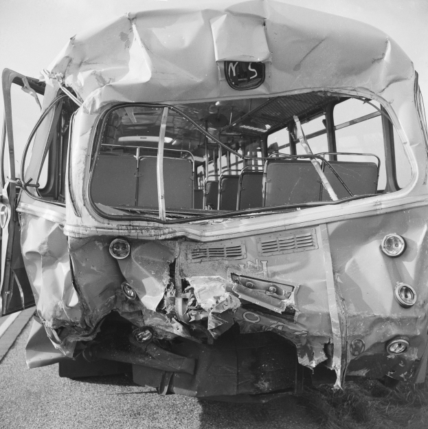 Ein Schwarz-Weiß-Foto eines schwer beschädigten Busses am Straßenrand, dessen Vorderseite eingedrückt und dessen Fenster zersplittert sind, mit der Straße und dem Himmel im Hintergrund.