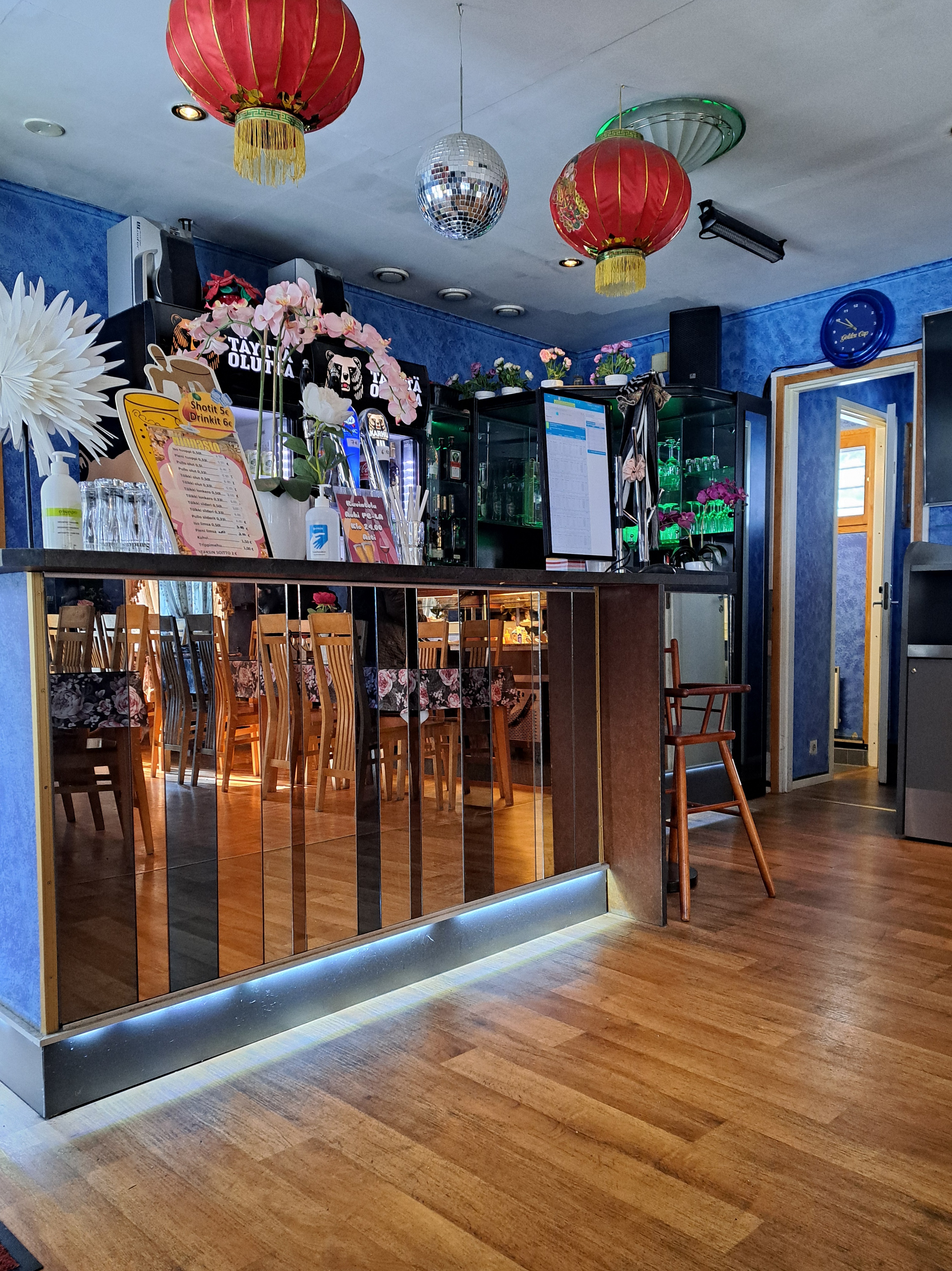 Das Innere eines Restaurants mit blauen Wänden und roten Laternen an der Decke, das Tische und Stühle, einen zentralen Schreibtisch mit Schränken, Tafeln mit Text, Blumenvasen, Dekorationsgegenstände, eine Tür und eine Wanduhr zeigt.