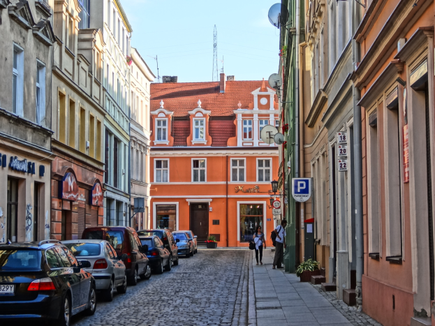 Eine Kopfsteinpflasterstraße in Prag, Tschechische Republik, mit geparkten Autos, Fußgängern, Gebäuden mit Fenstern, Topfpflanzen, Schildern, Satellitenschüsseln und einem Turm im Hintergrund unter dem Himmel.