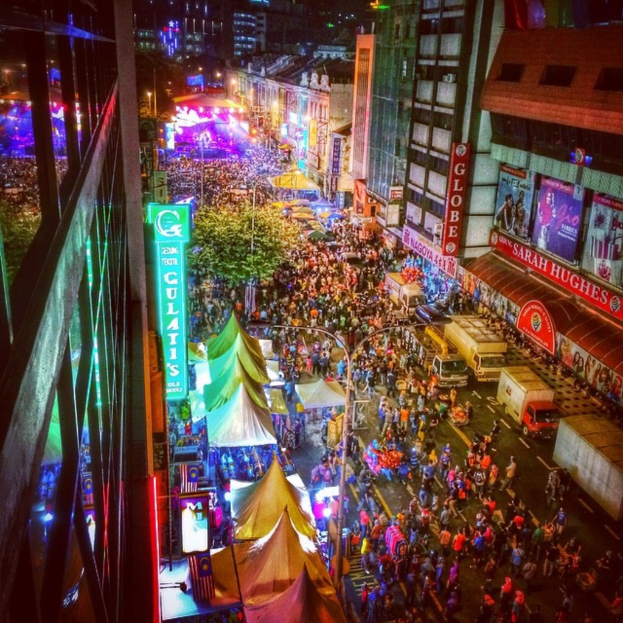 Eine belebte Volksfestszene mit einer großen Menschenmenge auf der Straße, umgeben von Zelten, Geschäften, Gebäuden und Bannern, mit zahlreichen Lichtern, die den Hintergrundbereich erhellen.