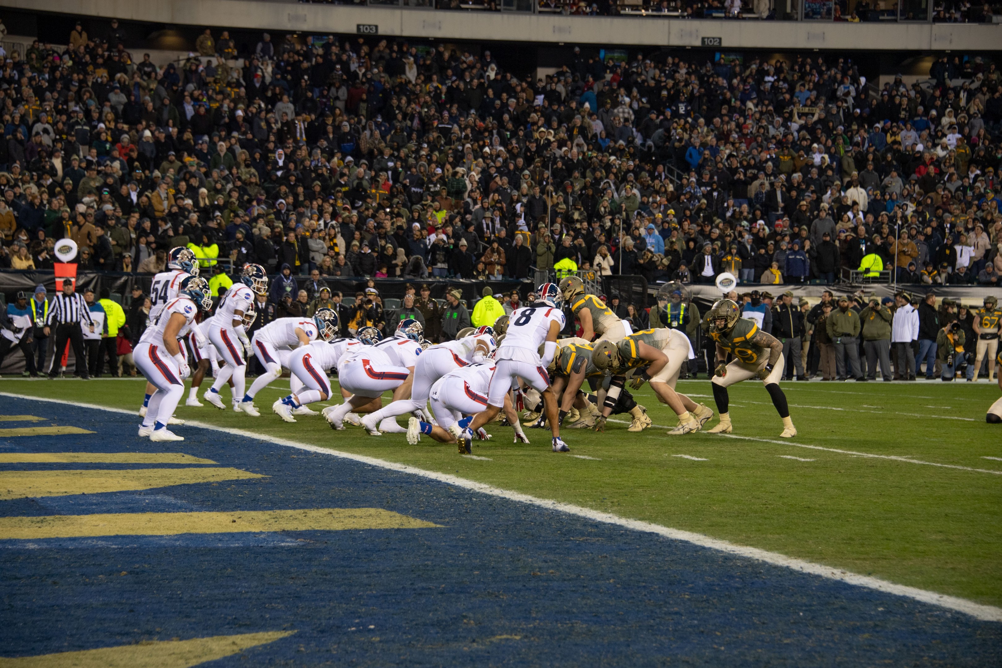 Ein Fußballspiel im Gange mit Spielern in Sportbekleidung und Helmen auf dem Boden, von einem Stadionplatz aus mit Zuschauern, Geländern und Beschilderung im Hintergrund betrachtet.