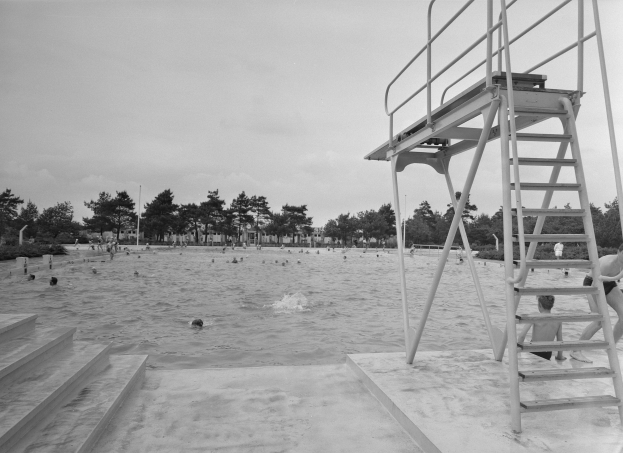 Ein Schwarz-Weiß-Foto von Menschen, die im Wasser an einem Strand schwimmen, mit einem Lebensrettungsturm auf der rechten Seite mit Stufen, die hinaufführen, und Bäumen, Pfählen und einem klaren Himmel im Hintergrund.
