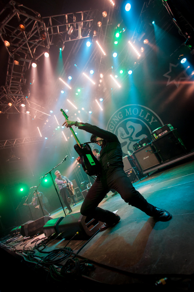 Ein musikalisches Konzert mit drei Künstlern auf der Bühne, zwei spielen Gitarre mit Mikrofonen davor, ein Hintergrund mit dem Wort "Molly" und zwei Schlangensymbole, sowie Scheinwerfer darüber.