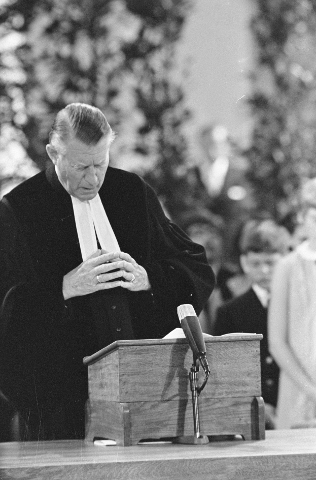 Ein Mann in einem schwarzen und weißen Gewand steht an einem Rednerpult mit einem Mikrofon, umgeben von ein paar Menschen und Bäumen im Hintergrund.