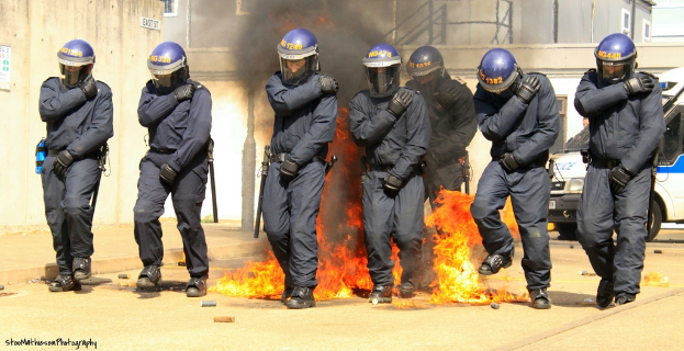 Männer in Helmen vor einem Feuer mit Gegenständen auf dem Boden, Gebäuden im Hintergrund, einem Fahrzeug und einem Plakat und einer Tafel an der linken Wand, mit Text am unteren Rand des Bildes.