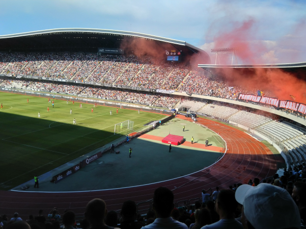 Ein großes Stadion voller Zuschauer bei einem Fußballspiel, mit Spielern im Einsatz auf dem Feld und Text am unteren Bildrand.