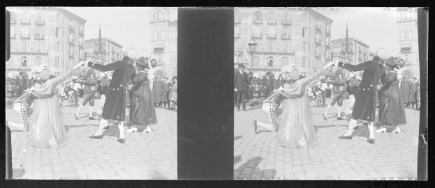 Ein Schwarz-Weiß-Foto einer Gruppe von Menschen in Kostümen, die auf der Straße tanzen, umgeben von Gebäuden, Straßenlaternen und einer klaren Himmel.