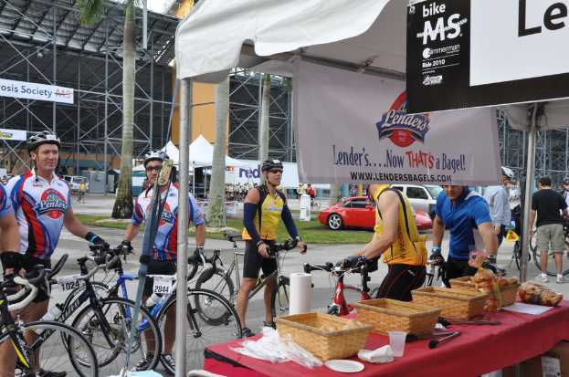 Eine Straßenszene mit Menschen in der Nähe von Fahrrädern, ein Geschäft mit Gegenständen auf einem Tisch, Bäume, Zelte, an einer Hütte befestigte Banner, Fahrzeuge und Gras.