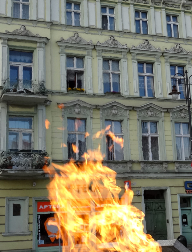 Ein großes Feuer brennt vor einem Gebäude mit Fenstern und Türen, einem Laternenmast und einer Tafel mit Text darauf.
