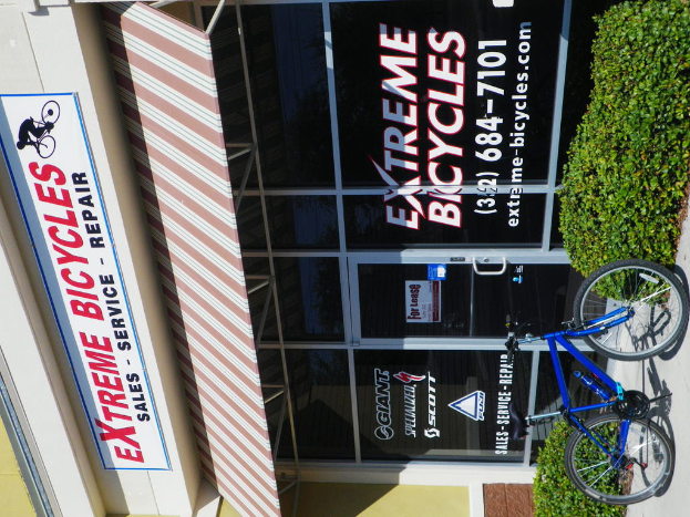 Ein Geschäft mit einer Glastür vorne, ein Schild links, ein geparktes Fahrrad rechts und kleine Pflanzen in der Nähe.