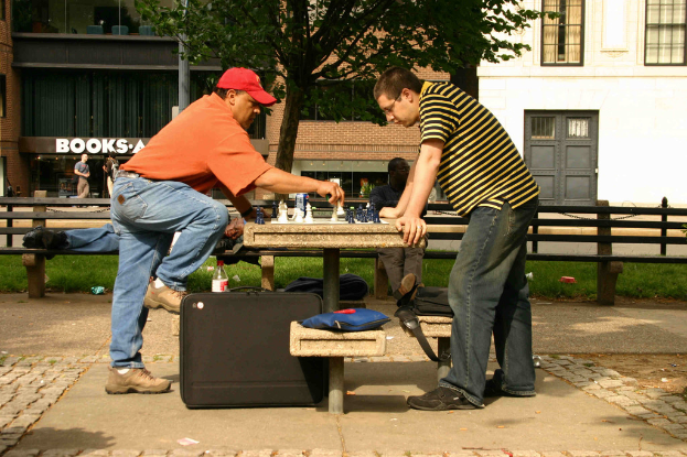 Ein Mann mit rotem Hut sitzt an einem Tisch mit einem Schachbrett und Figuren, ein großer Koffer daneben, während ein anderer Mann auf einer Bank hinter ihm schläft, mit einem Gebäude, Baum und Gras im Hintergrund.