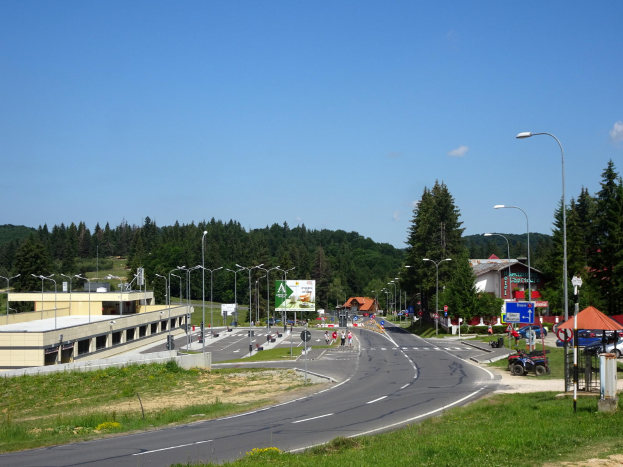 Ein Kreisverkehr mit Fahrzeugen, Gras, Laternenmasten, Texttafeln, Gebäuden, Bäumen und einem bewölkten Himmel, mit einem Straßenschild im Vordergrund.