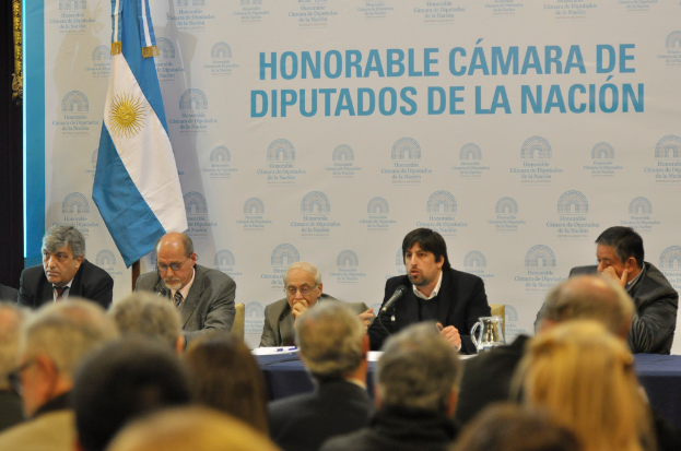 Eine Gruppe von Menschen sitzt an einem Tisch mit einem Krug, Papieren und einem Stift, während eine Person in der Mitte in ein Mikrofon spricht, mit einer weißen und blauen Flagge und einem Hintergrund, auf dem 'ehrenwerte kamara de' steht.