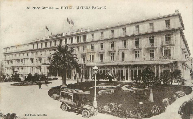 Eine schwarz-weiße Postkarte des Hotels Riviera-Palace in Nice-Cimiez mit einem Auto, Menschen und dem Gebäude mit Fenstern, Bäumen, Pflanzen, Laternen und Fahnen mit Stangen.