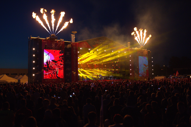 Große Menschenmenge auf einem Musikfestival mit beleuchteter Bühne, umgeben von Bannern, Zelten links und Feuerwerk am dunklen Himmel.