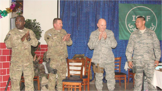 Vier Personen stehen, drei klatschen, eine sitzt auf einem Stuhl neben einer Pflanze, mit einem Banner an einem Vorhang im Hintergrund, alle tragen militärische Kleidung, mit Stühlen und einem Tisch im Sichtfeld.
