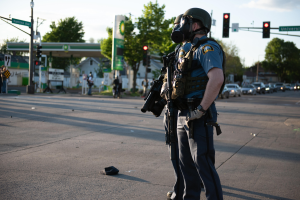 Ein Polizist in voller Ausrüstung, einschließlich einer Gasmaske, steht auf der Seite einer Straße und hält eine Waffe, mit Fahrzeugen, Verkehrszeichen, Schildern, Bäumen, Gebäuden und einer Tankstelle im Hintergrund.