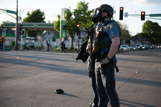 Ein Polizist in voller Ausrüstung, einschließlich einer Gasmaske, steht auf der Seite einer Straße und hält eine Waffe, mit Fahrzeugen, Verkehrszeichen, Schildern, Bäumen, Gebäuden und einer Tankstelle im Hintergrund.
