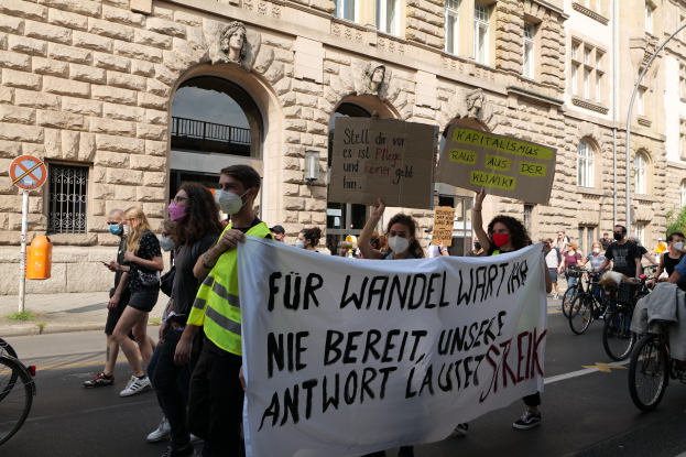 Eine Gruppe von Menschen marschiert auf der Straße in Berlin, hält Schilder und Banner hoch, während einige Fahrräder fahren, vor einem Gebäude mit Bögen, Säulen, Skulpturen und Bäumen.