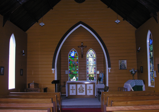Das Innere einer Kirche mit Bänken auf beiden Seiten, einem Kreuz in der Mitte, hängenden Lampen oben, einem Tisch mit einer Vase und einem Rahmen auf der rechten Seite und einem zentralen Idol, flankiert von einem Tisch mit einer Decke.