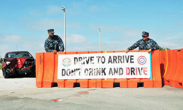 Zwei Polizisten neben einem beschädigten Auto mit einem Banner davor, mit Laternenmasten, Bäumen und einem klaren blauen Himmel im Hintergrund.