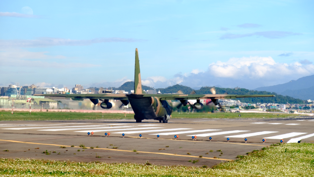 Ein großes Militärflugzeug startet von einer Flughafenrollbahn mit Gras und kleinen Pflanzen im Vordergrund, Gebäuden, Bäumen, Bergen und einem klaren blauen Himmel im Hintergrund.