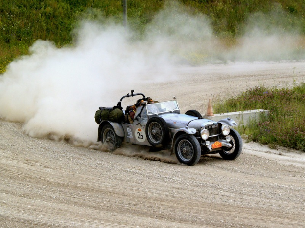 Ein Mann fährt ein Auto auf einer Schotterstraße mit Rauch aus dem Auspuff, umgeben von Pflanzen und Bäumen, mit einem Verkehrskegel im Vordergrund.