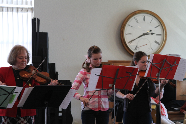Drei Frauen, die auf der Bühne Geigen spielen, während das Publikum zuhört, mit Notenpulten vor ihnen, einer Uhr an der Wand und einem weißen Vorhang im Hintergrund.
