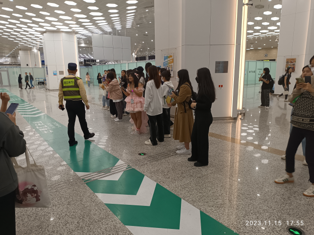 Eine Gruppe von Menschen steht in einer Reihe an einem Flughafen-Sicherheits-Check-in, einige halten Taschen und Mobiltelefone, mit Säulen im Hintergrund und Lampen an der Decke und einem Wasserzeichen am unteren Rand des Bildes.