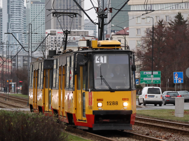 Eine gelbe und rote Straßenbahn f√§hrt auf Schienen neben hohen Geb√§uden, mit Stromm√§sten, Kabeln, Fahrzeugen, Schildern, B√§umen und einem klaren blauen Himmel.