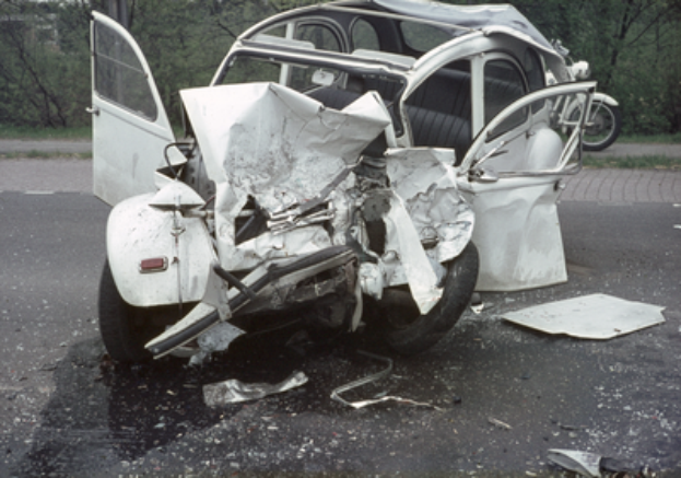 Ein verunfalltes Auto mit schwerer Frontalbeschädigung am Straßenrand, mit Bäumen, einem Gebäude und einem anderen Fahrzeug im Hintergrund.