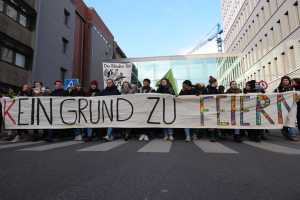 Eine Gruppe von Menschen auf der Straße mit einem Banner, auf dem "Kein Grund zu Feiern" steht, vor Gebäuden, einem Schild, einem weiteren Banner, einem Kran und einem bewölkten Himmel.