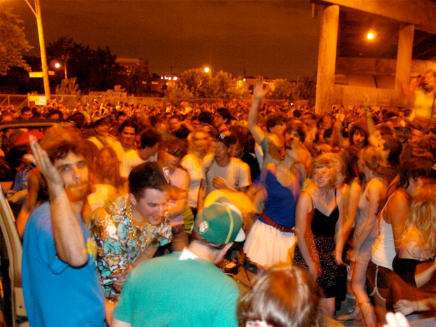 Menschen tanzen im Vordergrund eines Konzerts mit Lichtern, Pfählen, Bäumen und anderen Objekten im Hintergrund.