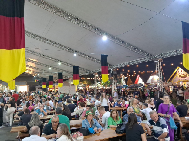 Eine große Gruppe von Menschen sitzt an Tischen unter einem Zelt beim Oktoberfest in München, Deutschland, mit einigen Stehenden und Taschen auf den Tischen, umgeben von Zelten, Lichtern und Fahnen.