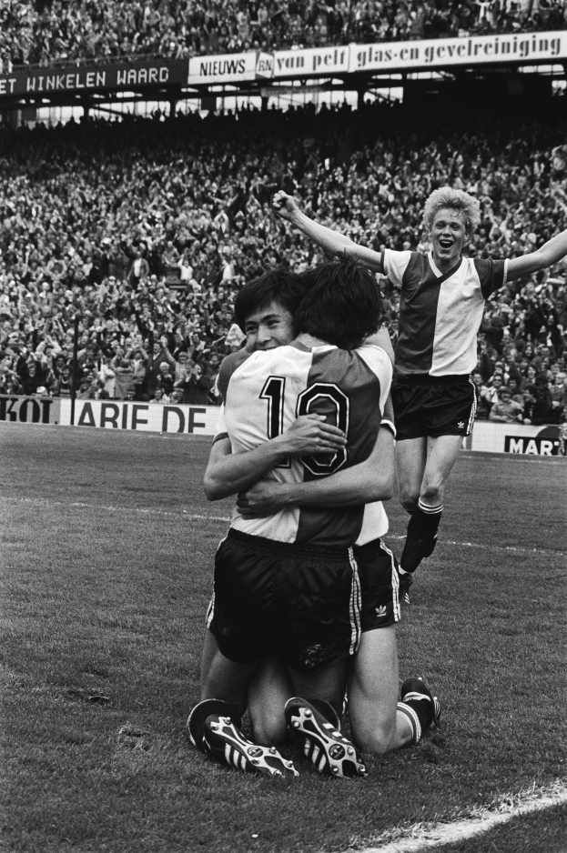 Drei Fussballspieler umarmen sich auf dem Boden, feiernd nach einem Tor vor einer jubelnden Menge in einer schwarz-weißen Stadionszene.