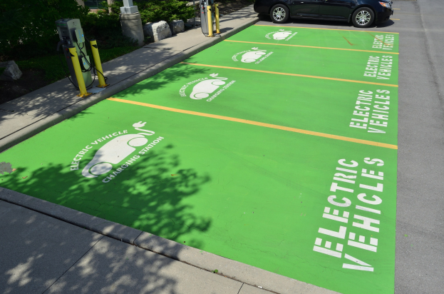 Elektroauto auf einer Ladestation mit grüner Bodenmatte mit Text und Bildern in einer Parkanlage umgeben von Bäumen, Pflanzen, Pfählen und anderen Gegenständen.