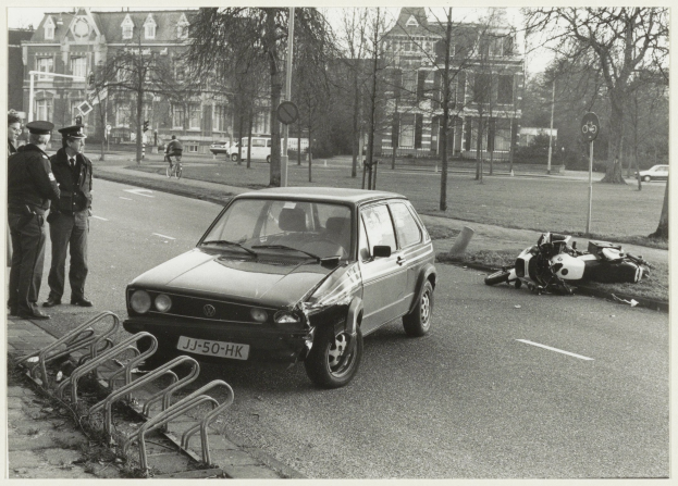 Ein Schwarz-Weiß-Foto eines Autos, das auf der Straße in ein Motorrad gekracht ist, mit drei Personen auf dem Gehweg links neben dem Auto und Bäumen, Pfählen, Gebäuden und anderen Fahrzeugen im Hintergrund.