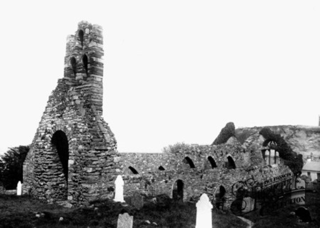 Alte Steinkirche in Ruinen mit einem Friedhof im Vordergrund und einem Gebäude im Hintergrund, unter einem sichtbaren Himmel mit verstreuten Bäumen.