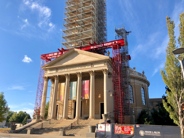 Ein große Baustelle mit Gerüten auf dem Dach, mit Pfeilern, Treppen mit Geländern, Bannern mit Text, einem Laternenmast, Bäumen und Fahrzeugen auf der Straße im Vordergrund, mit einem bewölktem Himmel im Hintergrund.