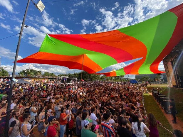 Eine große Menge Menschen steht vor einer Regenbogenfahne, mit einem Gebäude, einer Bühne, Lautsprechern, Bäumen und Wolken im Hintergrund bei einer Festival-Jubiläumsfeier.