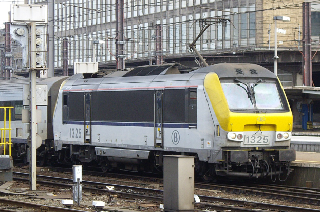 Ein dieselhydraulischer Lokomotive fährt auf Schienen neben einem hohen Gebäude, mit Strommasten und Drähten daneben, einer Plattform auf der rechten Seite und einem klaren blauen Himmel im Hintergrund.