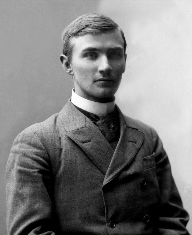 Schwarzes und weißes Porträt eines Mannes in Anzug und Krawatte, der mit gefalteten Händen und ernstem Gesichtsausdruck vor der Kamera steht.