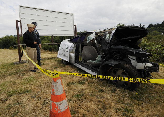 Ein Polizeibeamter in Uniform steht neben einem schwerbeschädigten Auto am Straßenrand, mit eingedellter Haube und zersplitterten Fenstern, neben einem Verkehrskegel und einem bandagierten Pfahl, mit Bäumen und einem bewölkten Himmel im Hintergrund.