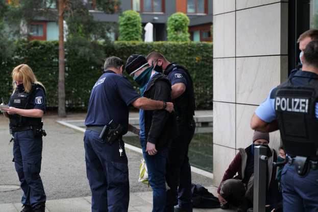 Eine Gruppe von Polizisten steht vor einem Gebäude, eine Frau hält ein Buch und einen Stift in der Hand, während einige Menschen in der Nähe stehen und sitzen und Pflanzen, Bäume und Gebäude im Hintergrund zu sehen sind.