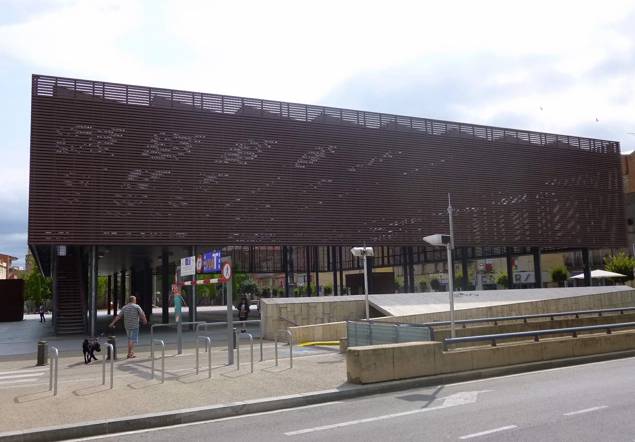 Großes modernes Gebäude mit zahlreichen Fenstern, das als neuer Eingang zur Stadt dient, flankiert von Straßeninfrastruktur und Fußgängern.